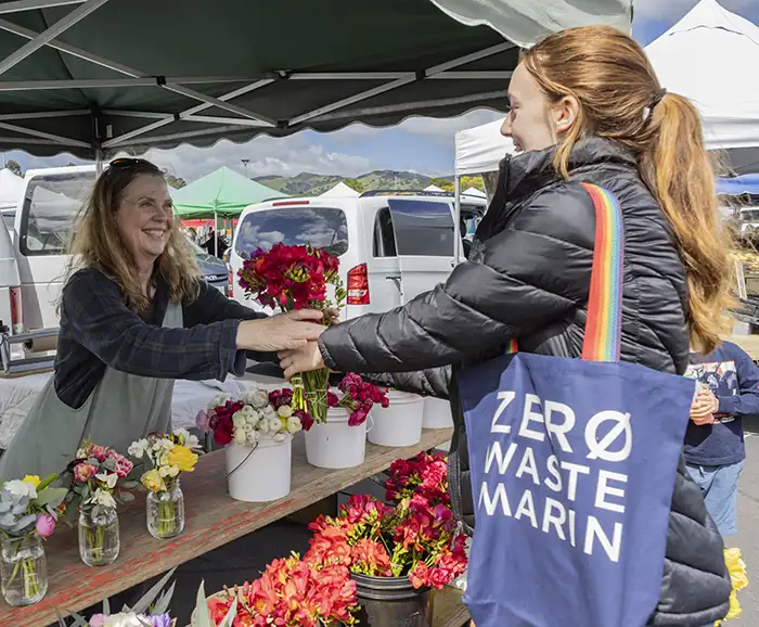 A woman with a Zero Waste Marin reusable bag reaches for flowers from a vendor at the Marin Farmer's Market