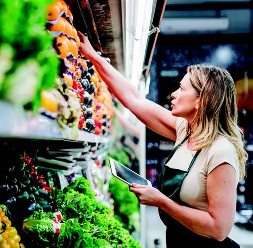 A woman in apron reaches for produce in a supermarket aisle