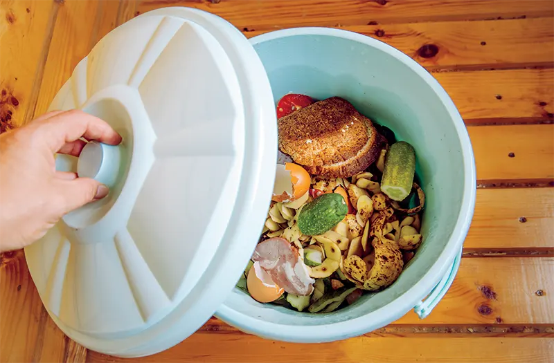 A kitchen compost pail with food scraps in it on a wood backdrop
