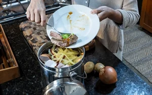 A person scrapes a plate of food scraps into a kitchen compost pail